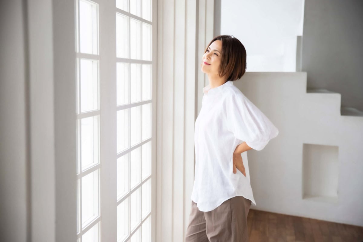 明るい窓の前でやわらかな表情を見せる、SHIGETA主宰のホリスティックビューティーコンサルタントCHICO SHIGETAさん。白いブラウスとブラウンのパンツ姿