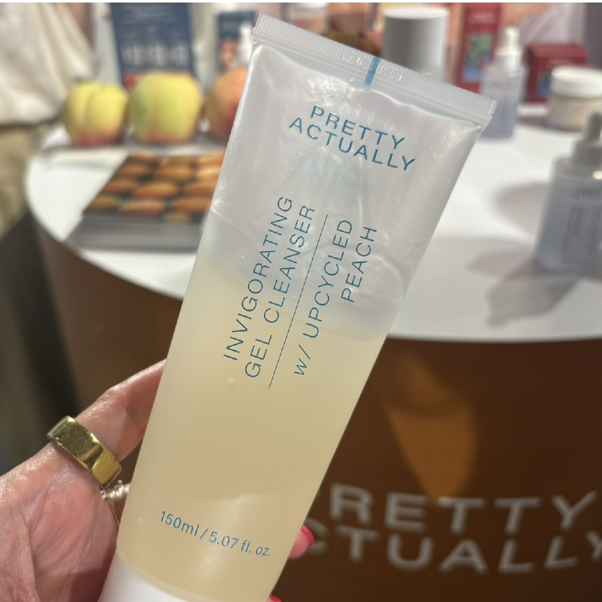 アップサイクルピーチを使用した「プリティアクチュアリー(Pretty Actually)」のジェルクレンザーのチューブ容器(150mL ¥2,000)。背景の展示台には、原料をイメージした本物の桃が置かれている。場所はコスメキッチン展示会。