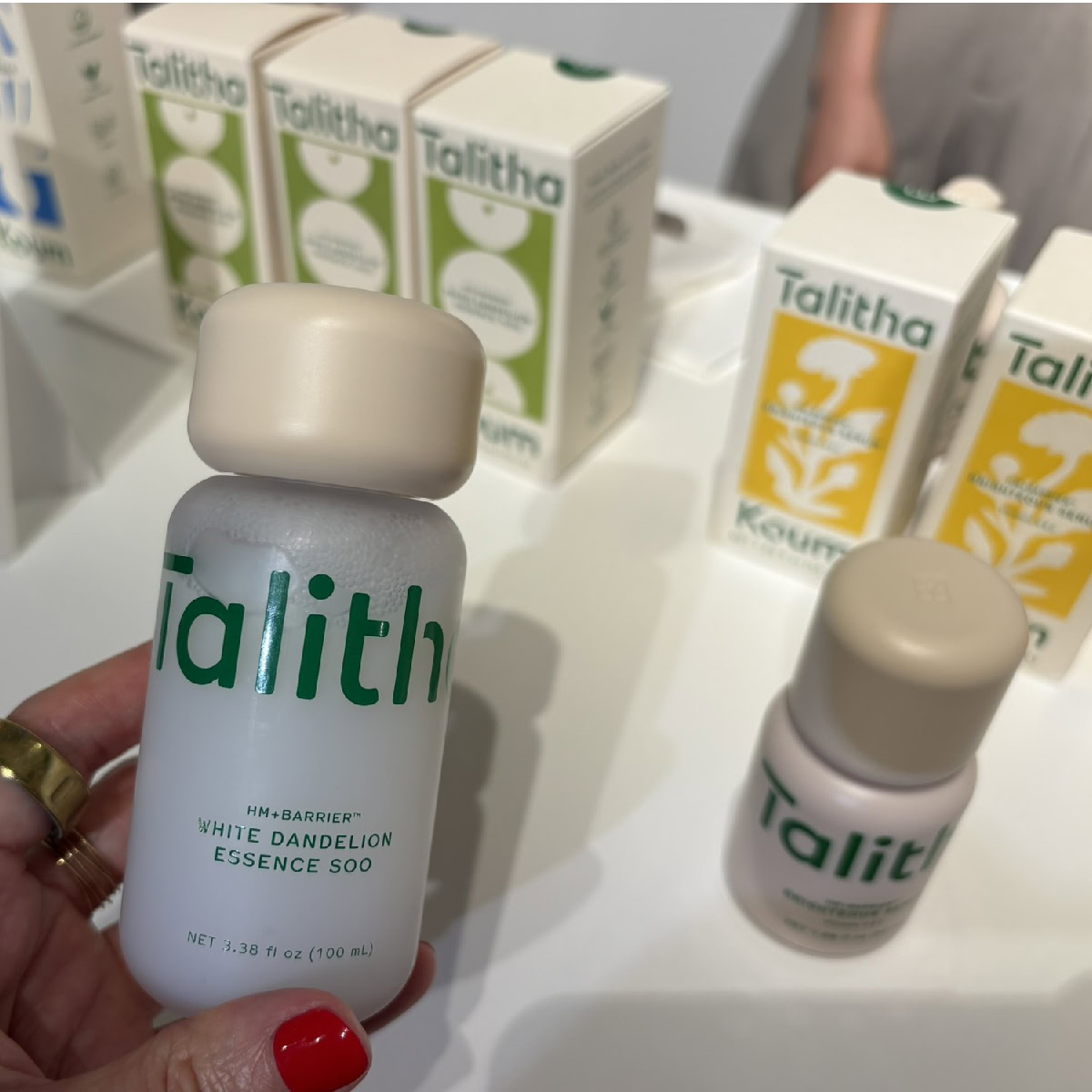 コスメキッチン展示会で撮影された、韓国コスメブランド「タリダクム(Talitha Koum)」のディスプレイ。手前には、ゴールドの指輪をはめ赤いネイルを施した女性の手に持たれた「白タンポポ エッセンスウォーター」の白いボトル(100mL ¥4,840)があり、奥には白と緑、白と黄色のパッケージが並ぶ。