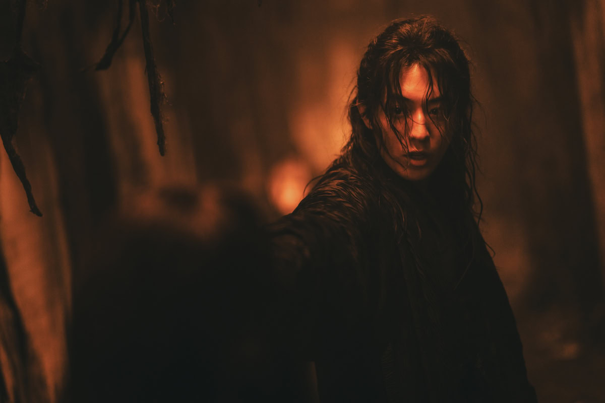 "Person with long wet hair in a dark, narrow, dimly lit corridor with an intense expression, reaching toward the camera, dramatic orange lighting creating a suspenseful and mysterious atmosphere."