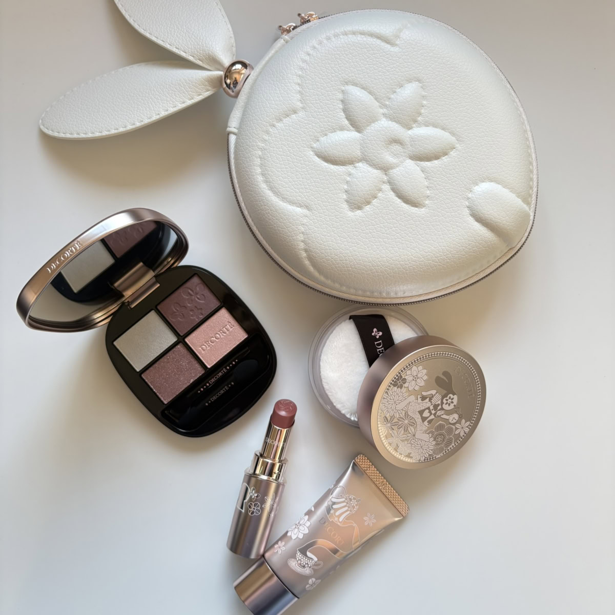 コスメデコルテの2026年ホリデー限定コフレ「メイクアップ コレクション 2026 Alice in DECORTÉ Wonderland」。白いうさぎ耳付き丸型ポーチ、4色アイシャドウパレット、リップスティック、化粧下地、フェイスパウダーの全5点が、白背景に平置きされています。