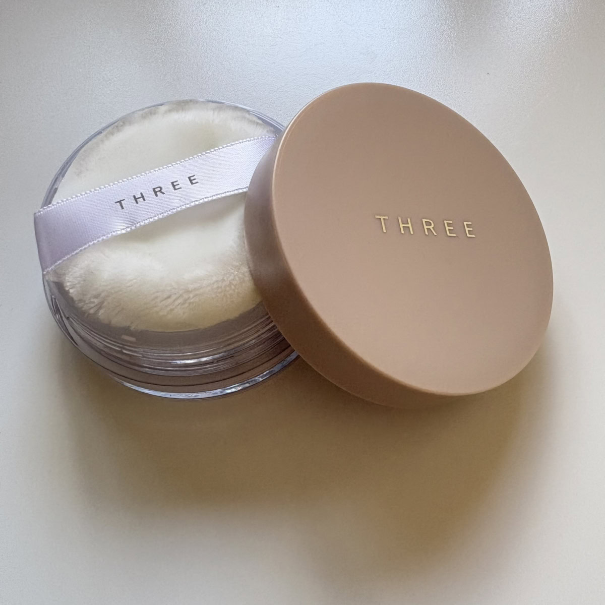 「THREE」のブランドロゴがゴールドで刻印されたベージュゴールドの蓋が、クリアな丸型容器の横に立てかけられた、「THREE クラウドヴェール ルースパウダー X02」の限定パッケージ。容器の中には、「THREE」のロゴ入り白いリボンが付いたふわふわとした白いフェイスパフが収められています。