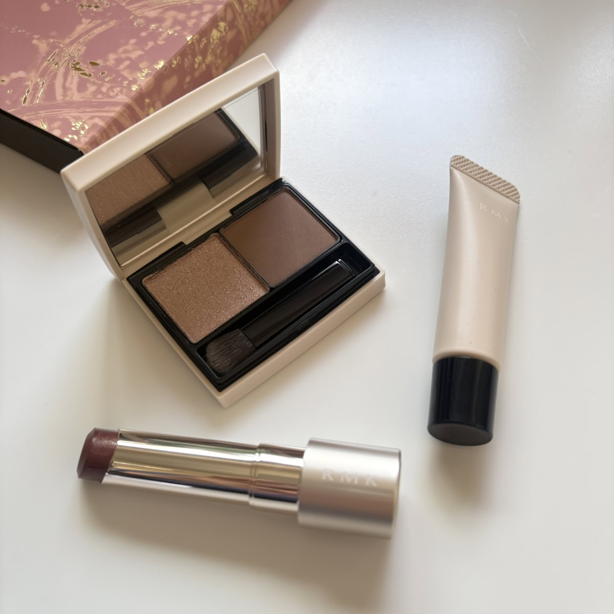 「RMK ザ クワイエット プロヴォカター キット」の全アイテムを上から撮影した写真。左上のピンクにゴールド柄の限定ボックスの手前に、開いた状態の2色アイシャドウパレット、その手前には、銀色のケースから少し繰り出したモーブトーンのリップスティック、右側には「RMK」ロゴが入った淡いベージュのチューブ型リキッドブラッシュが、白い背景に平置きされています。
