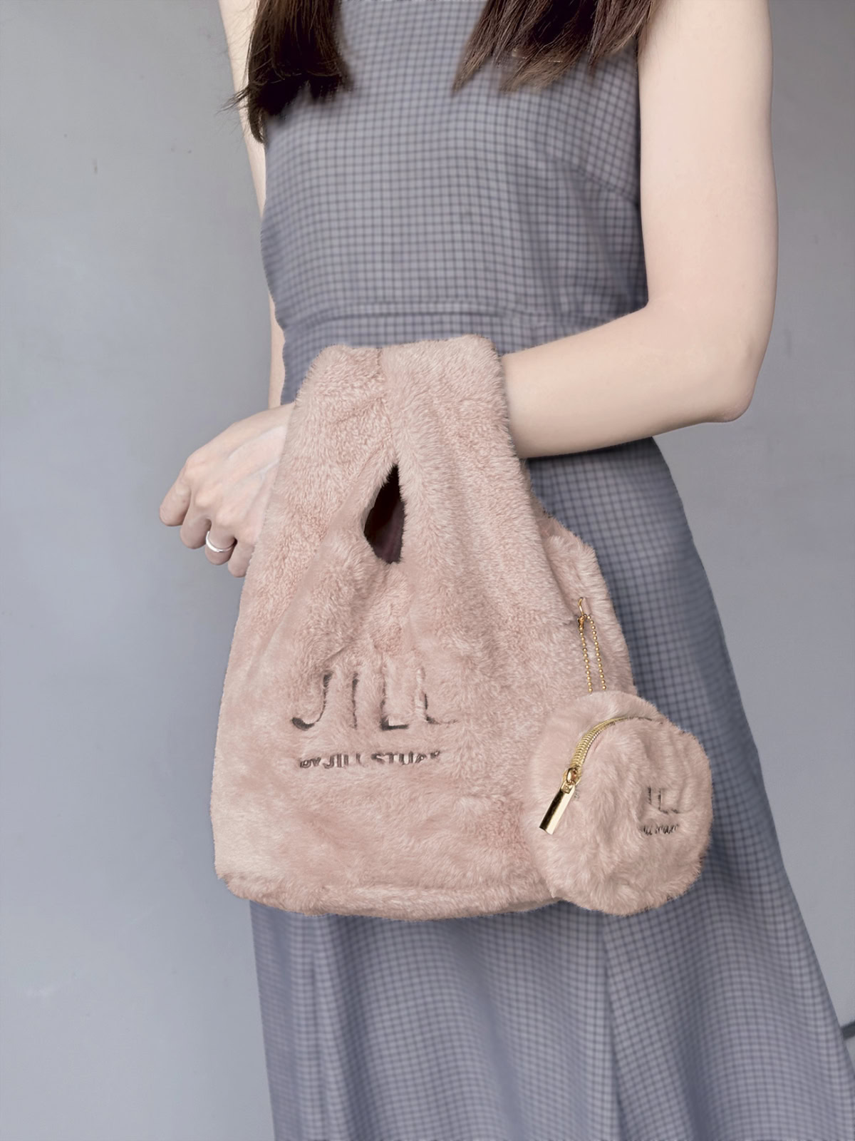 グレーのワンピースを着た女性が、腕に「JILL by JILL STUART」のロゴが入ったくすみピンクのふわふわファーバッグと丸型ポーチチャームを提げている着用イメージ。