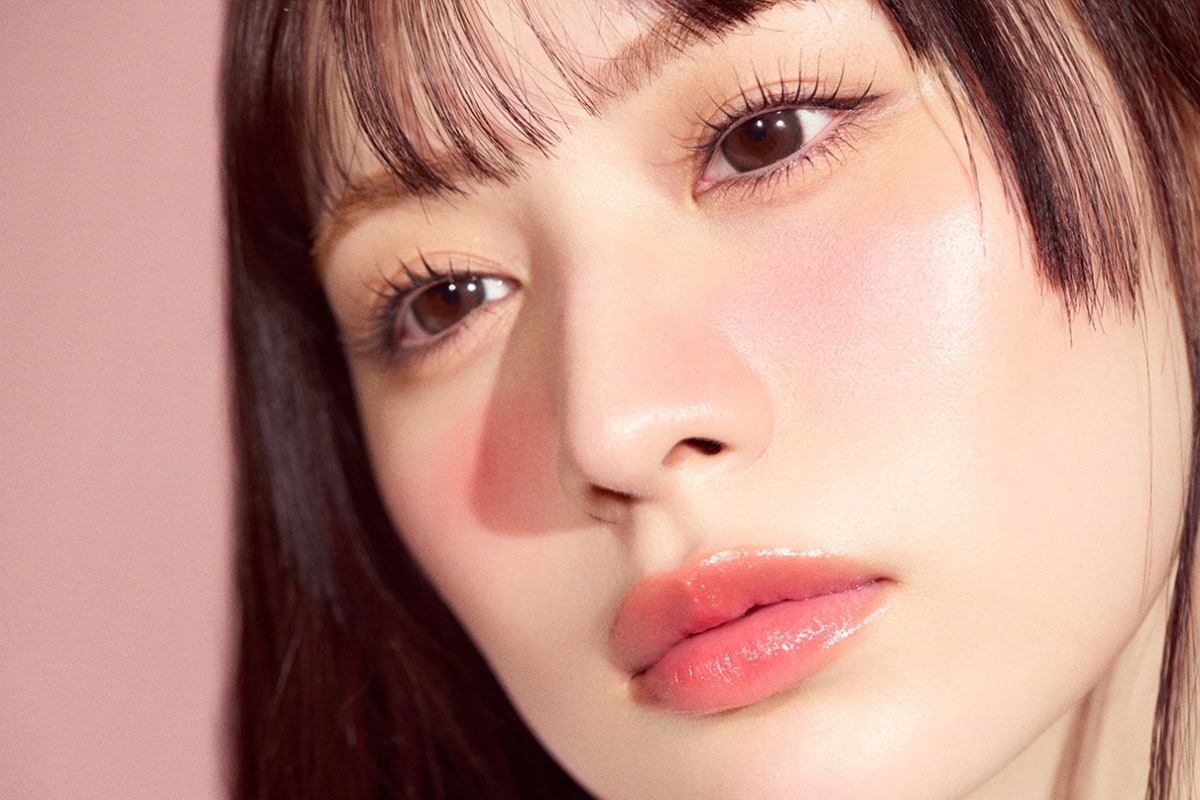 ピンクの背景で、伏し目でマスカラの仕上がりを見せる立花琴未のクローズアップ。