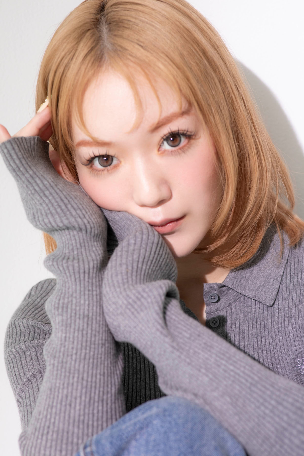 MA-さんの顔寄りのポートレート。明るいベージュカラーのボブヘアに、グレーのリブニットトップスを合わせている。袖に顔を添えながらカメラをまっすぐ見つめ、やわらかな自然光の中で印象的な表情を見せている。透明感のある肌と大きな瞳が目を引く一枚。