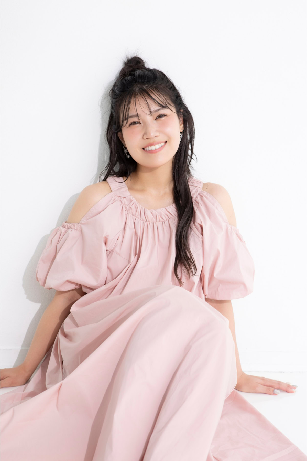 なるごりさんが淡いピンクのオフショルダーワンピースを着用し、白い背景の前に座って自然な笑顔を見せている写真。やわらかな自然光が差し込み、優しい雰囲気が印象的。