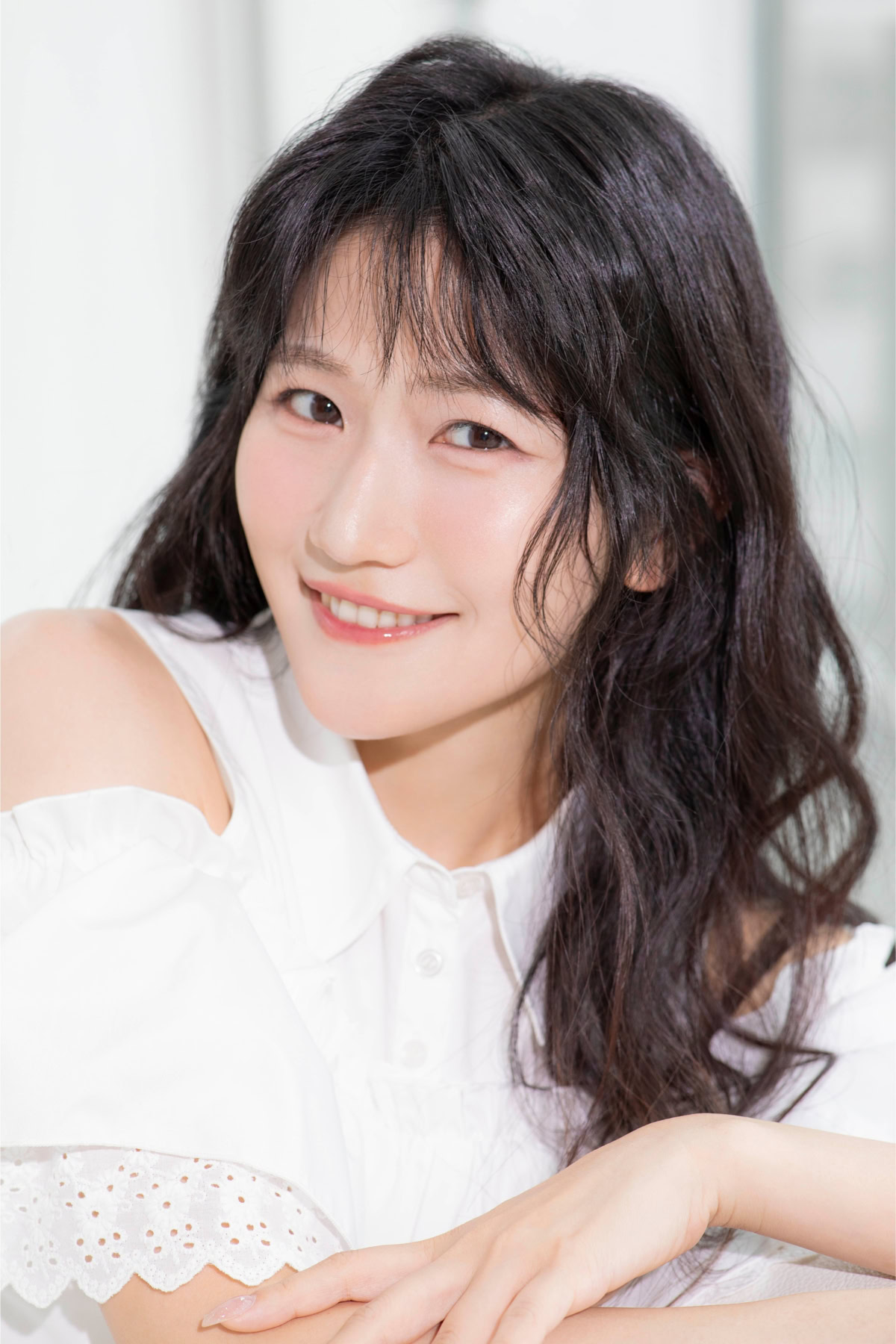 笑顔でカメラを見つめるるのるんさんの上半身ポートレート。白いオフショルダーデザインのブラウスを着用し、柔らかな自然光の中で腕を組みながらリラックスした表情を見せている。黒髪のゆるやかなウェーブと透明感のある雰囲気が印象的。