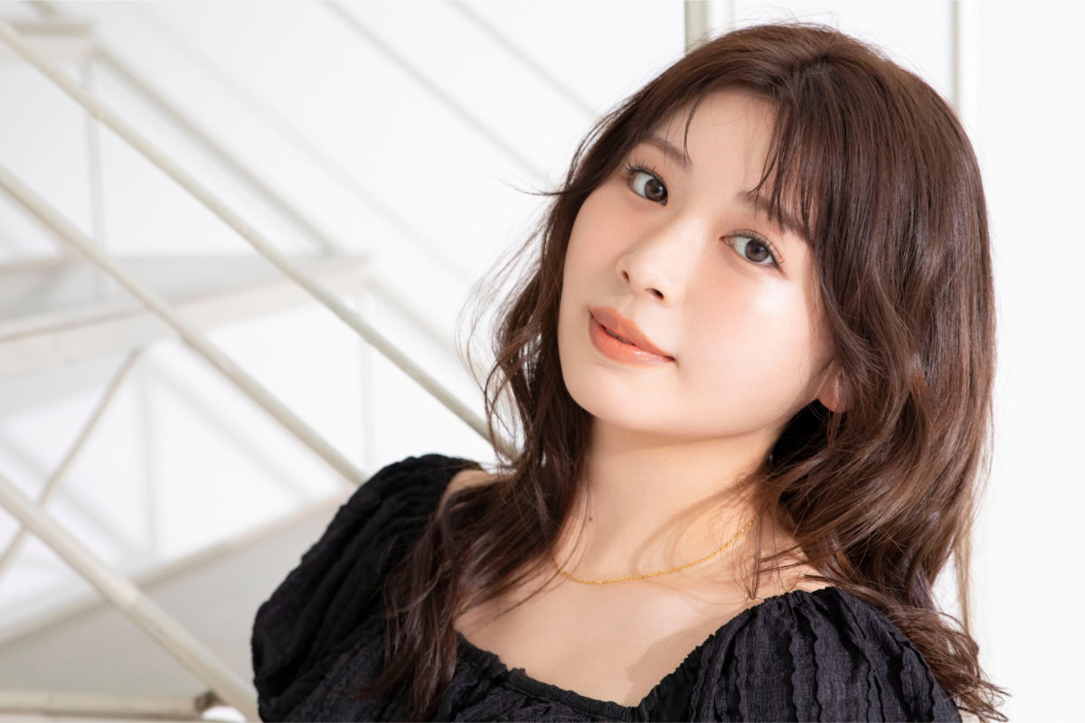 まるにゃさんの上半身ポートレート。黒のふんわりとしたトップスを着用し、やわらかな光が差し込む室内でカメラを見つめている。ゆるく巻かれたブラウンのロングヘアとナチュラルなメイクが上品な雰囲気を引き立てている。