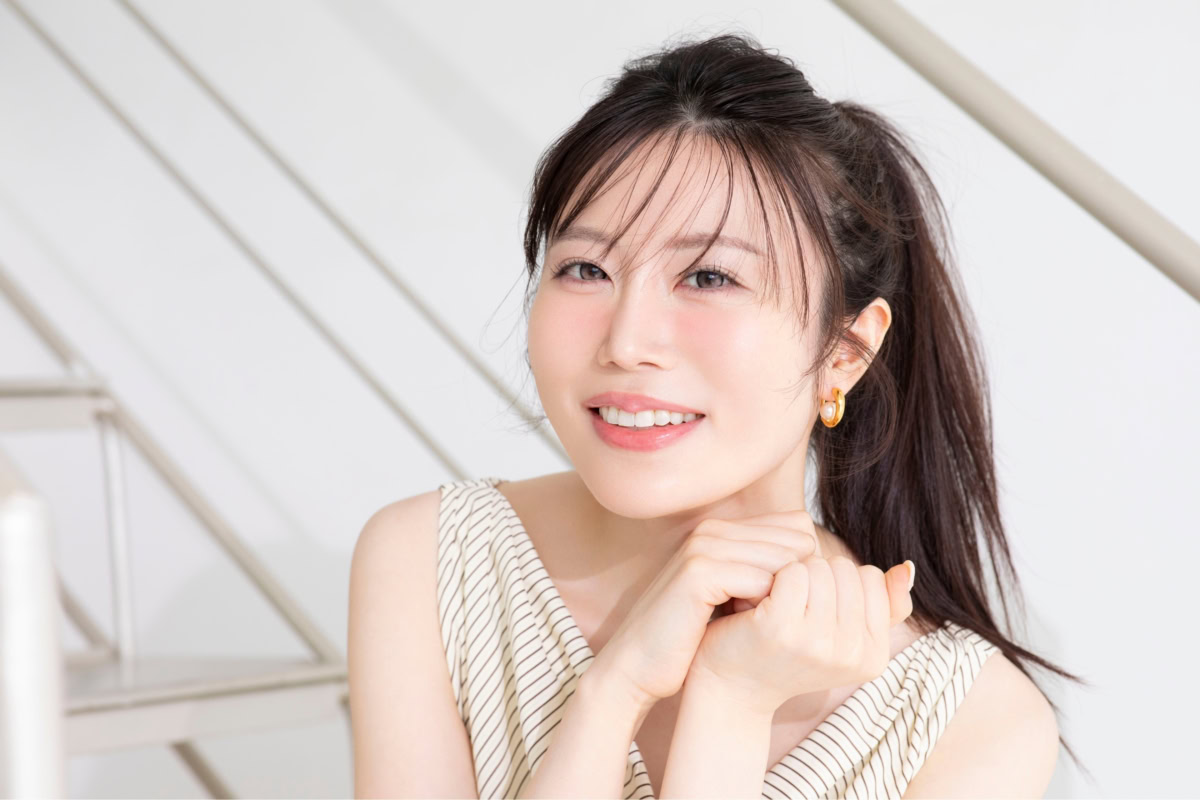 ときた総理さんの上半身ポートレート。ベージュ系のストライプ柄ノースリーブトップスを着用し、黒髪をポニーテールにまとめている。両手をあごの下で軽く組み、やわらかな自然光が差し込む室内でカメラに向かって爽やかな笑顔を見せている。