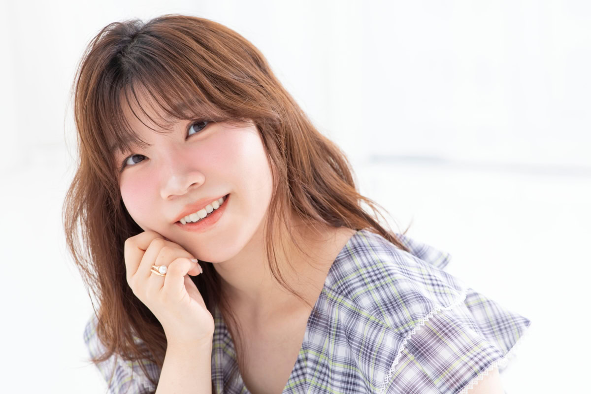 杏仁ゆんさんの上半身ポートレート。淡いパープル系のチェック柄トップスを着用し、頬に手を添えながらカメラに向かって明るく微笑んでいる。やわらかな自然光が差し込む室内で撮影され、ふんわりとしたブラウンヘアと親しみやすい表情が印象的な一枚。